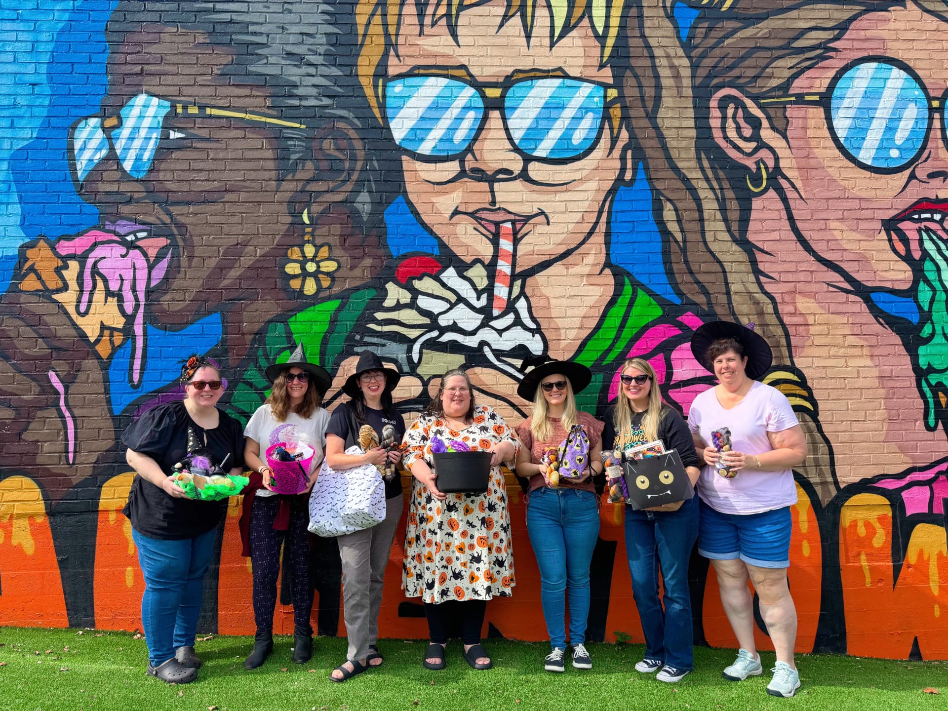 The Worsted Witches in witch hats posing with project bags in front of a colorful mural